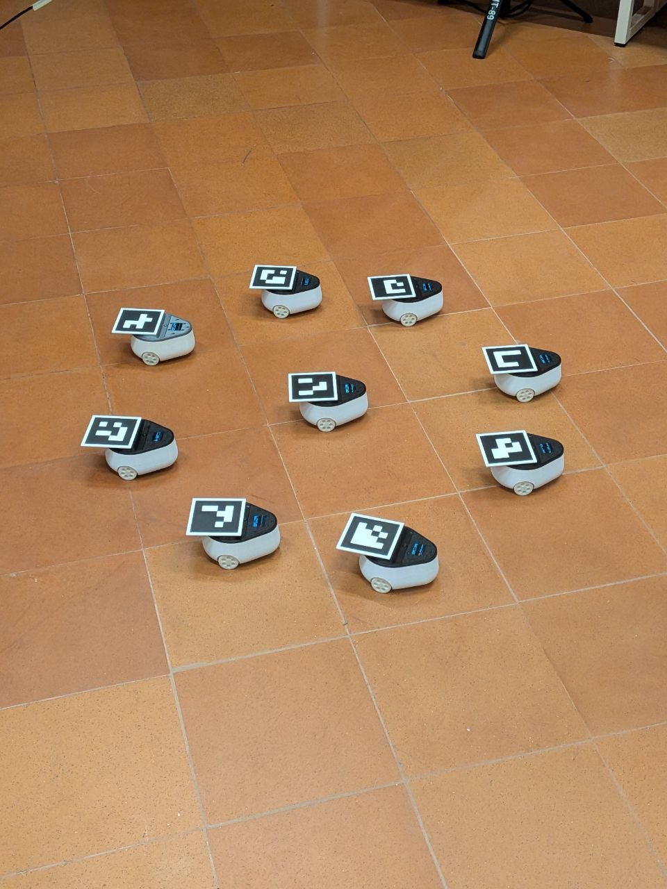 Nine small round white robots with ArUco markers arranged in a formation on a tiled floor.
