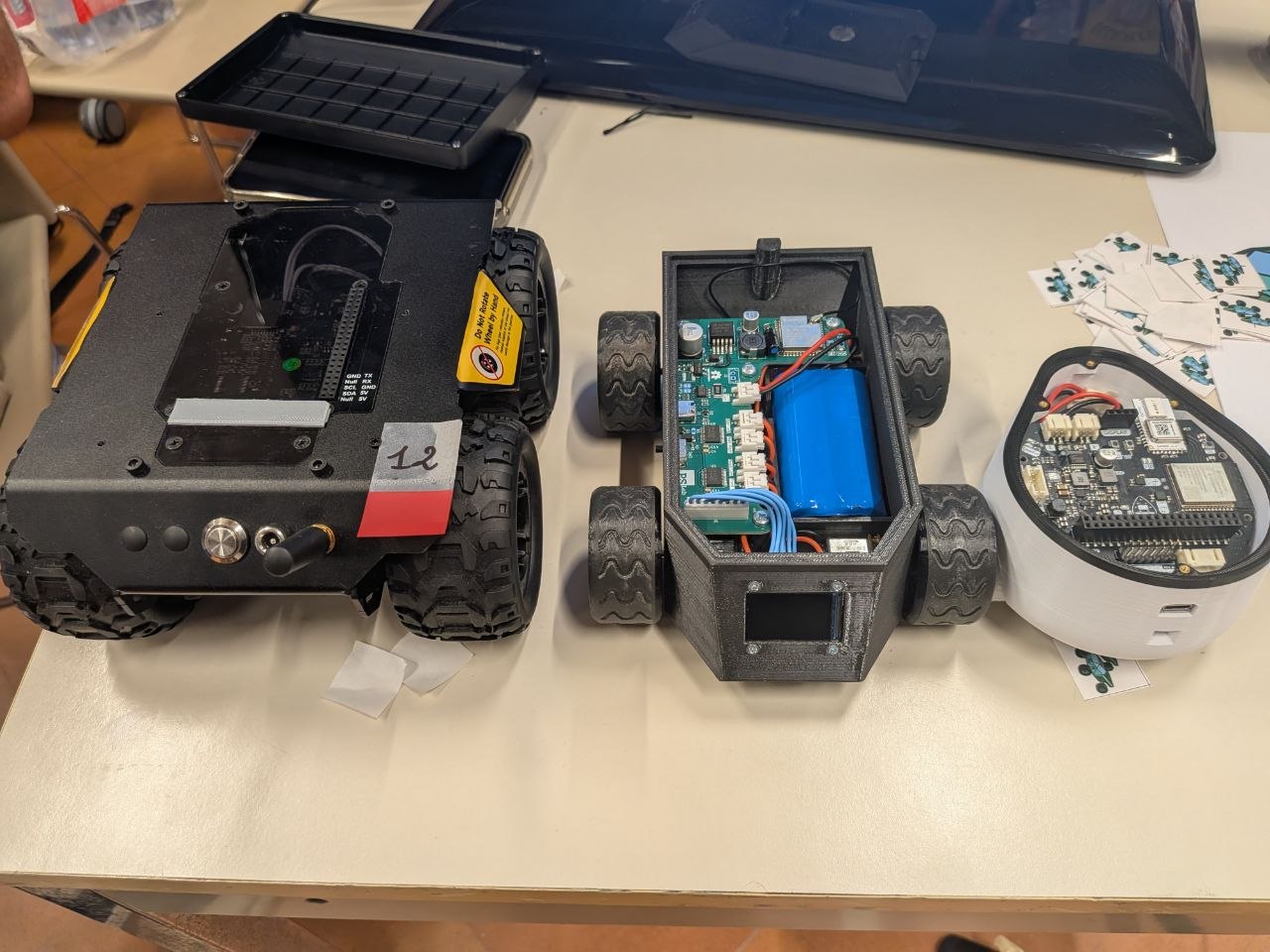 Three robots side by side on a table: a large black commercial rover, a rectangular 3D-printed robot with four wheels, and a small round white robot.