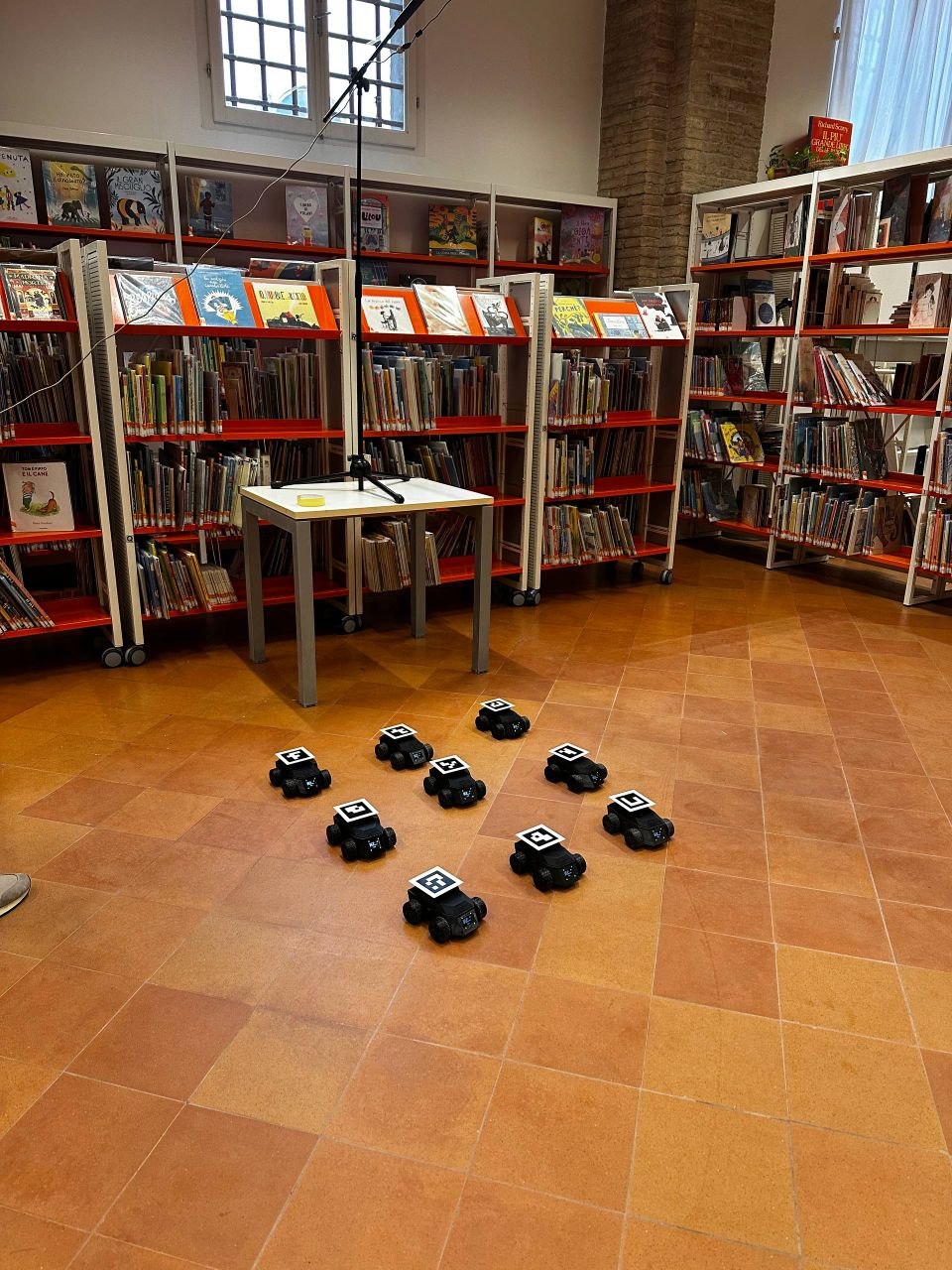 Nine black rectangular robots with ArUco markers on a tiled floor in a library.